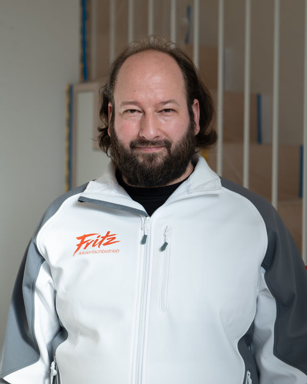 Volker, Teamleiter und Maler, bärtig in Arbeitsjacke des Fritz Malerfachbetriebs vor hellen Wandelementen