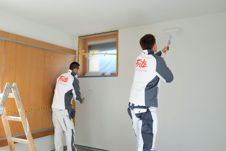 Zwei Maler in Fritz Malerbetrieb Arbeitskleidung streichen die Wände eines Raumes, Fenster und Boden sind abgeklebt